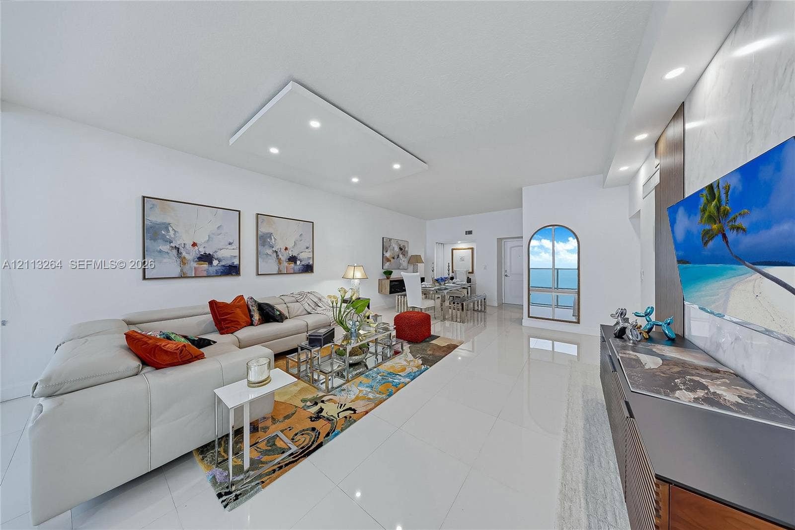 Open-concept living space featuring high-gloss tile flooring, recessed ceiling lighting, a custom stone-clad media wall, and an arched glass door