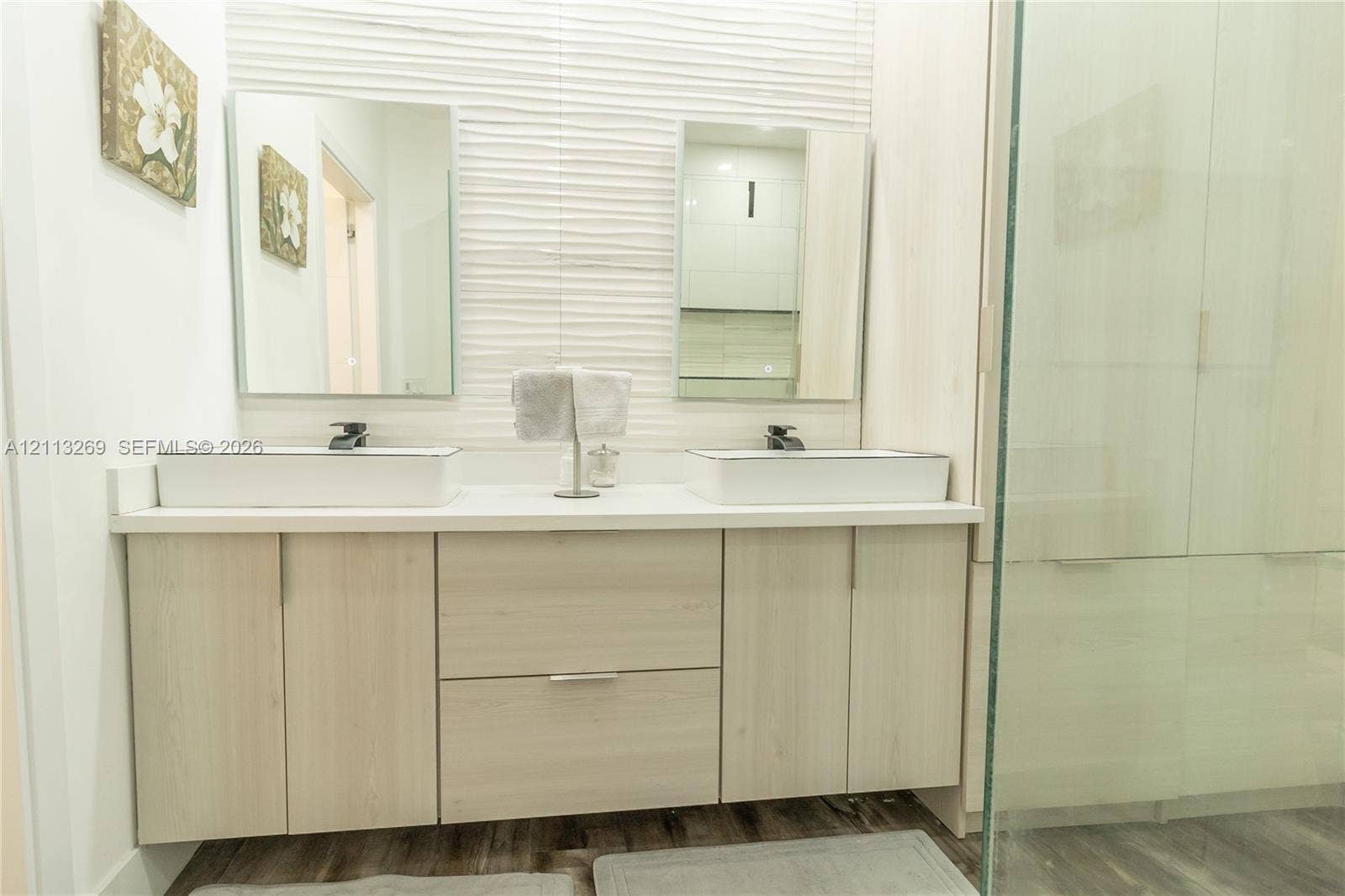 11494 NW 79th Lane, Doral, FL 33178 Double vanity featuring rectangular vessel sinks, light wood-tone cabinetry, a textured tiled wall accent, and frameless glass shower enclosure