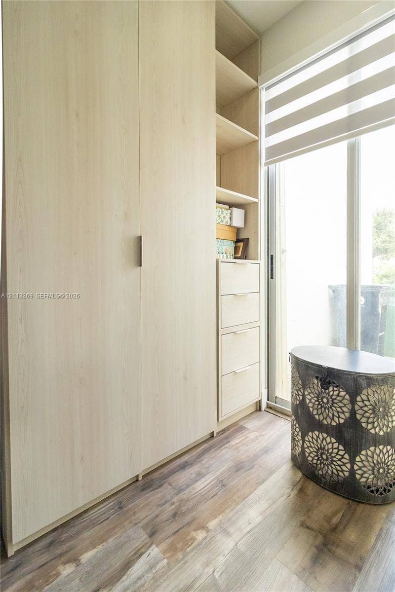 11494 NW 79th Lane, Doral, FL 33178 Light-toned wood cabinetry featuring integrated storage, open shelving, and drawer units with minimalist hardware, complemented by wood-finish flooring and a full-height glass door with installed roller shades
