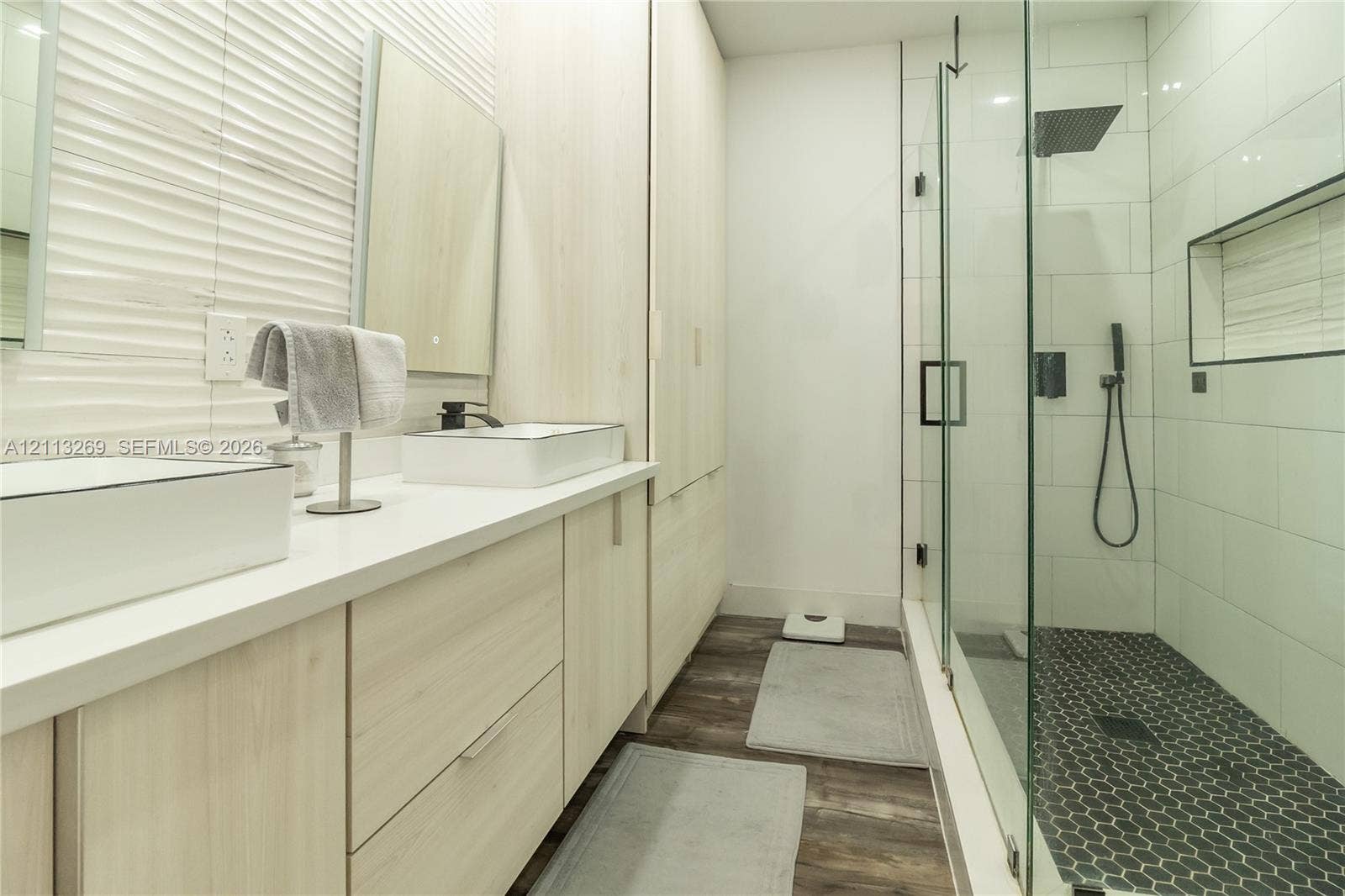 11494 NW 79th Lane, Doral, FL 33178 Dual-sink vanity with light wood-finish cabinetry, white solid-surface countertops, textured wall tile, frameless glass shower enclosure, and dark hexagon floor tile
