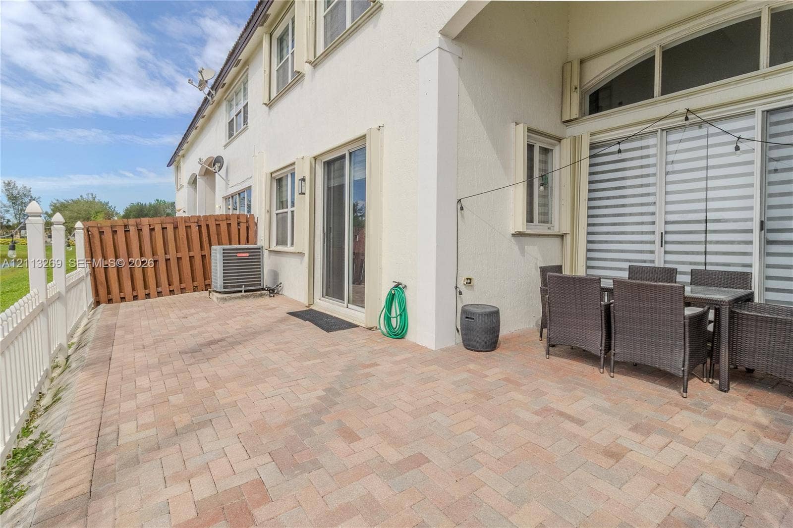 11494 NW 79th Lane, Doral, FL 33178 Rear patio featuring herringbone-patterned brick pavers, dual sliding glass door entry points, stucco exterior cladding, and white vinyl privacy fencing