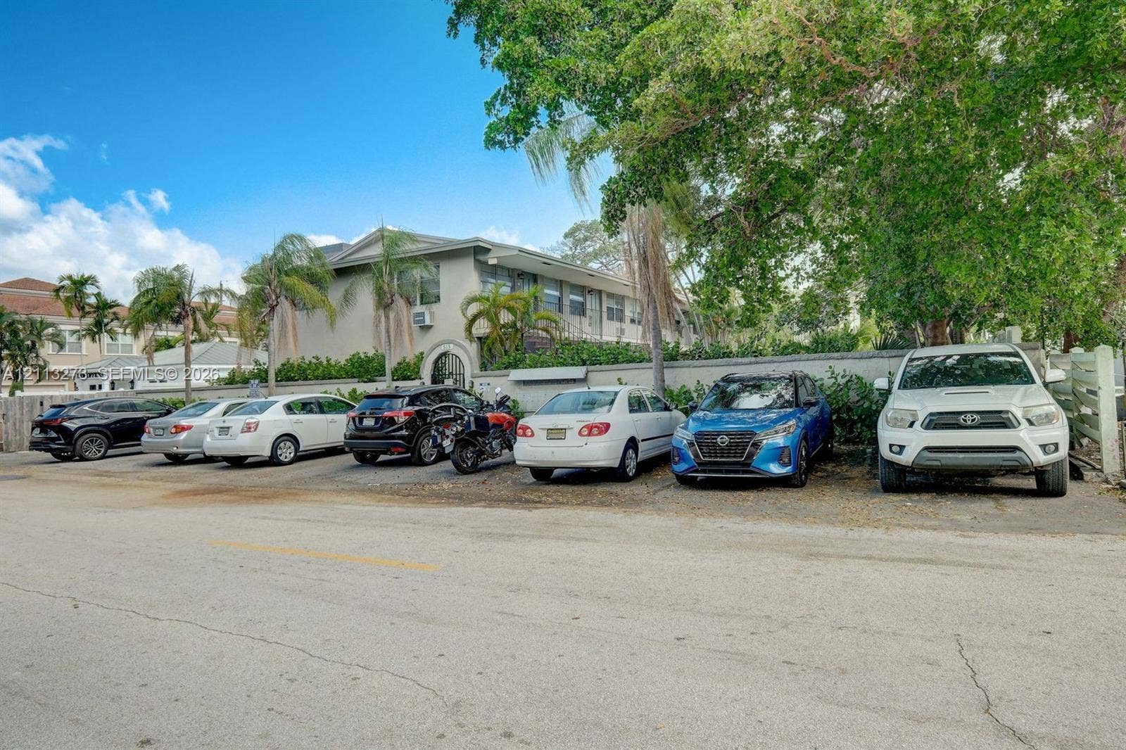 Multi-story exterior featuring smooth stucco siding, arched entryway, designated surface parking, and mature perimeter trees