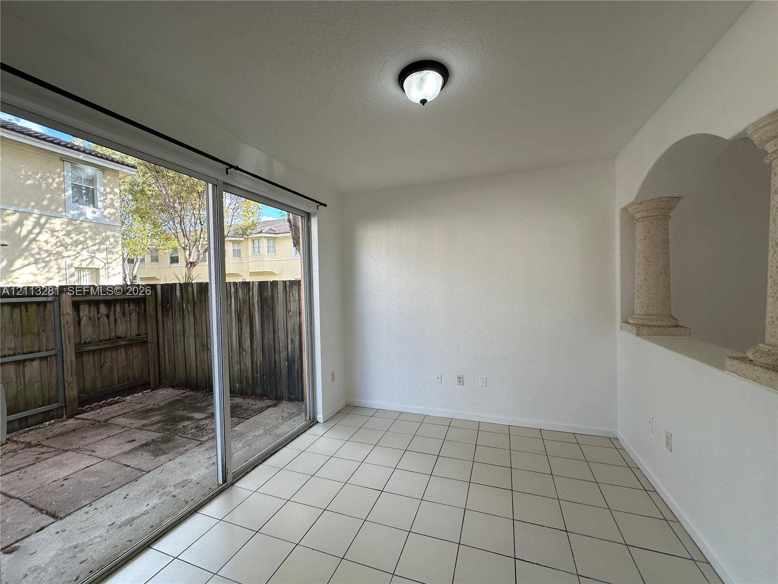 2615 SE 15th Place, #190, Homestead, FL 33035 Interior space featuring light-colored tile flooring, decorative architectural columns, a ceiling-mounted light fixture, and a large sliding glass door opening to a concrete patio enclosed by wood fencing