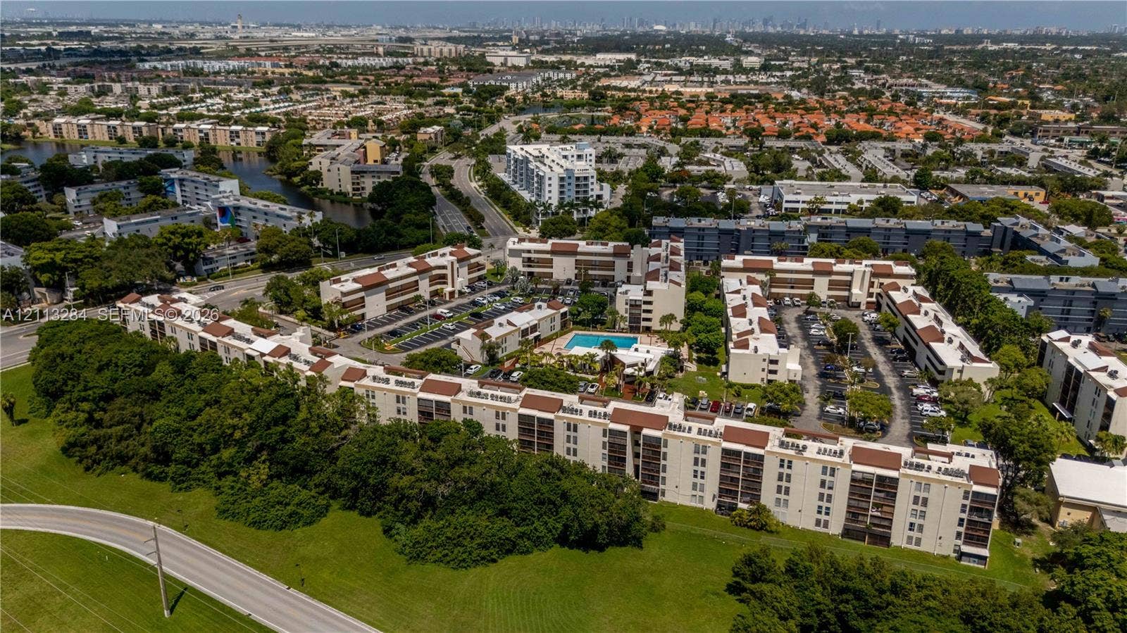 Aerial perspective of the condominium complex featuring multi-story structures with terra-cotta-toned roofing, an outdoor swimming pool, paved surface parking, and perimeter greenery