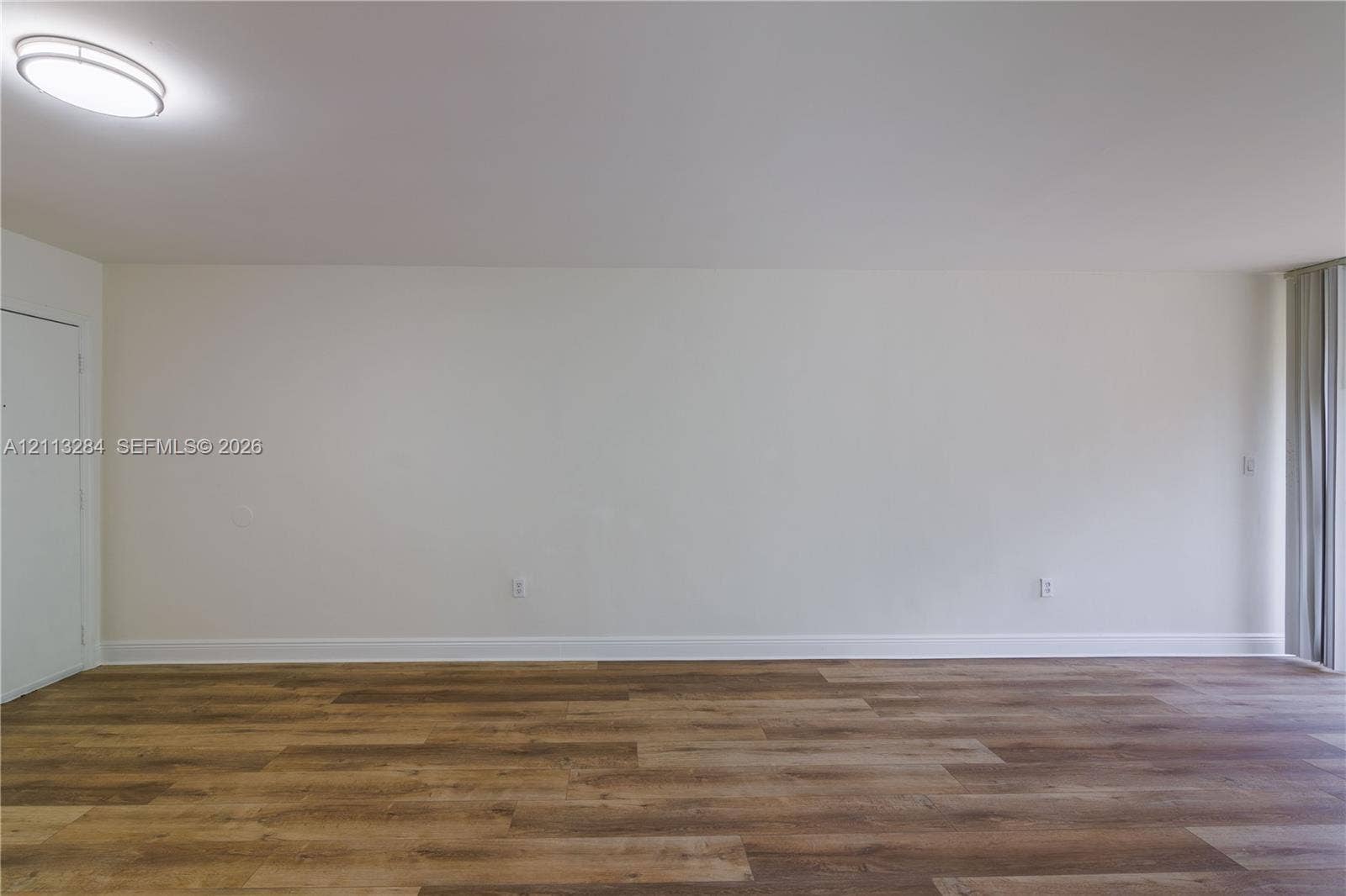 Interior space featuring wood-finish flooring, neutral wall tones, a ceiling-mounted flush light fixture, and vertical window blinds