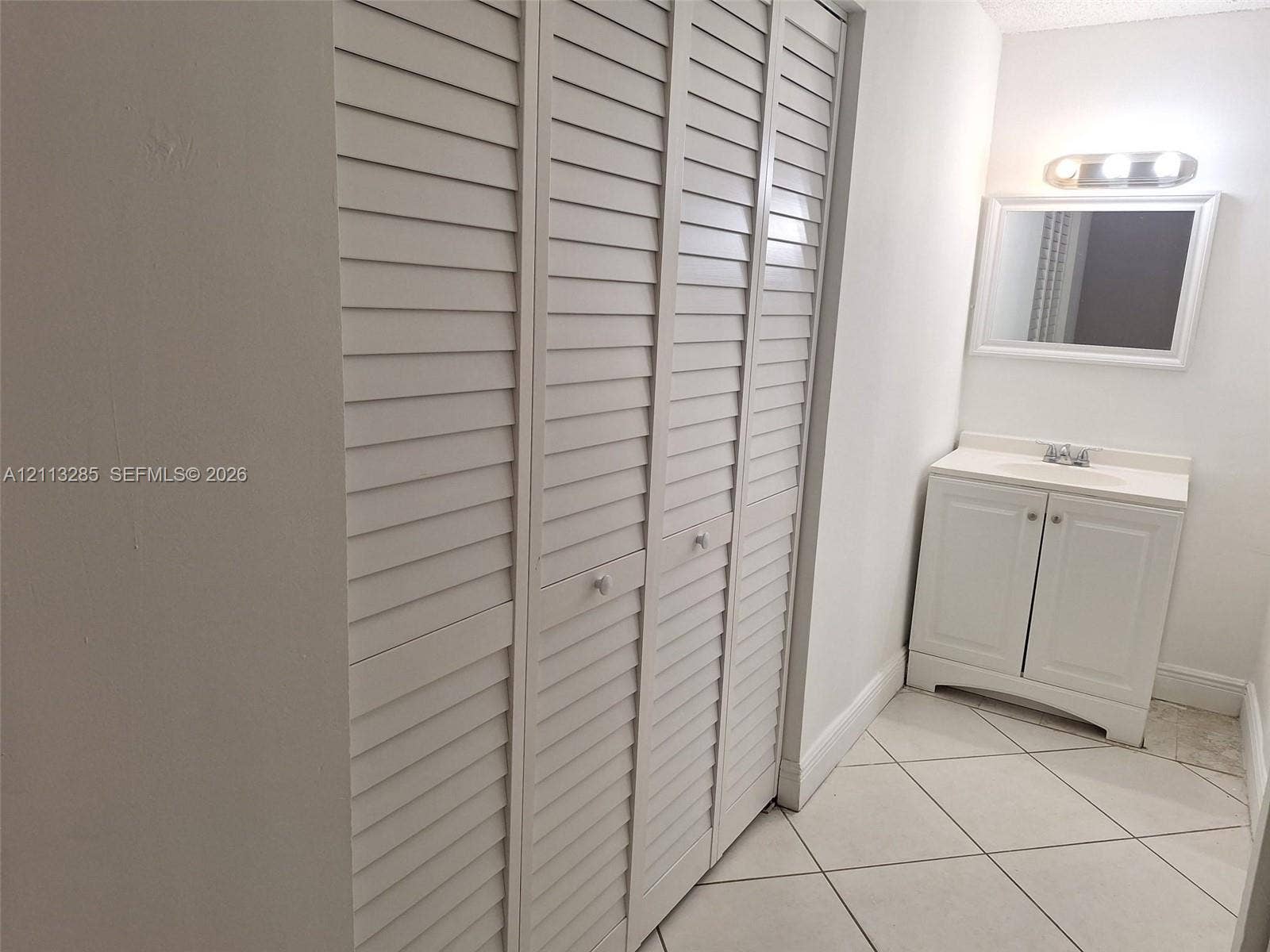 White louvered closet doors, vanity with integrated basin, polished light-colored tiled flooring, and wall-mounted vanity mirror with integrated lighting fixture