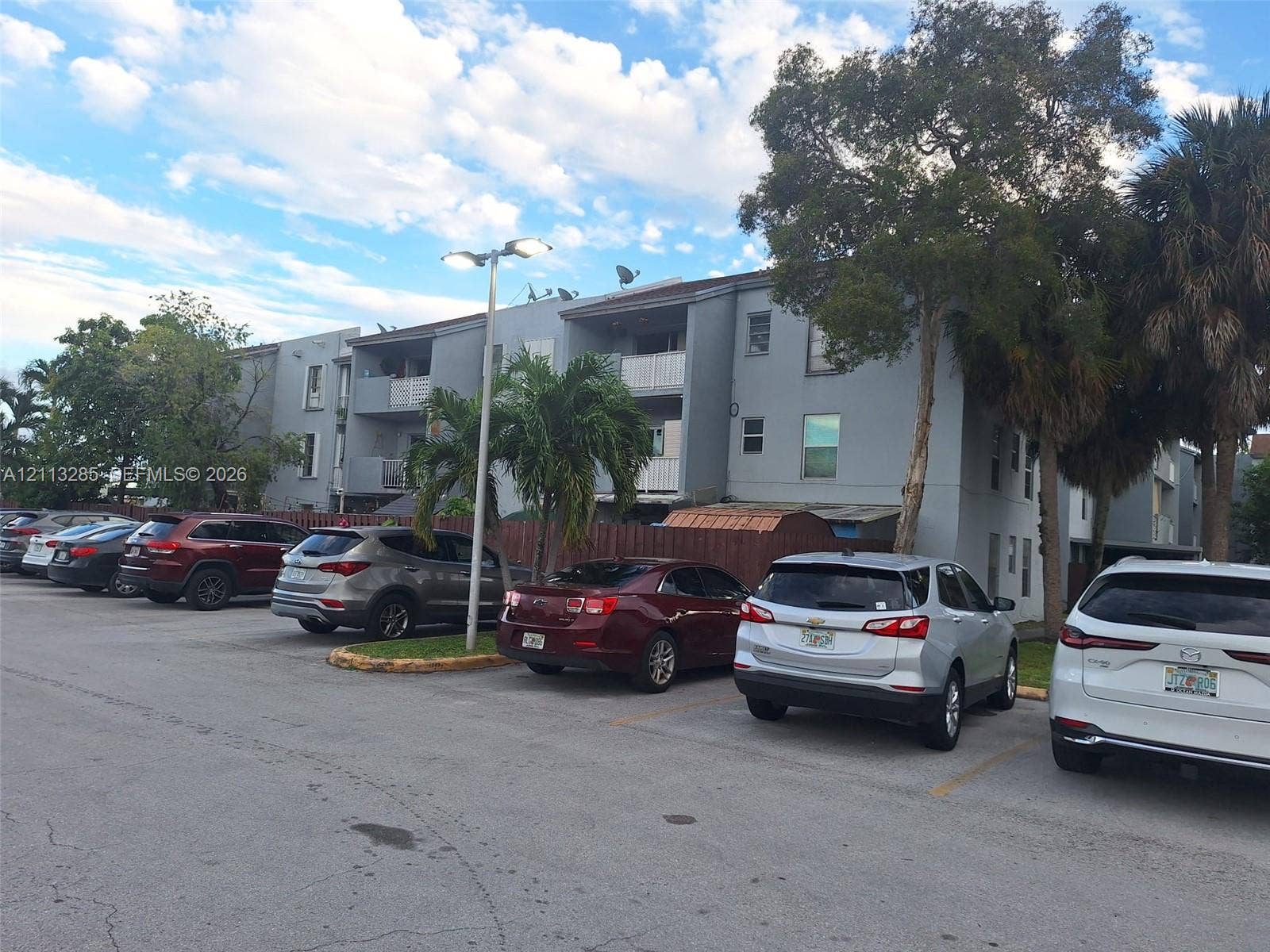 Multi-story building exterior featuring balconies, an asphalt parking lot, and mature trees