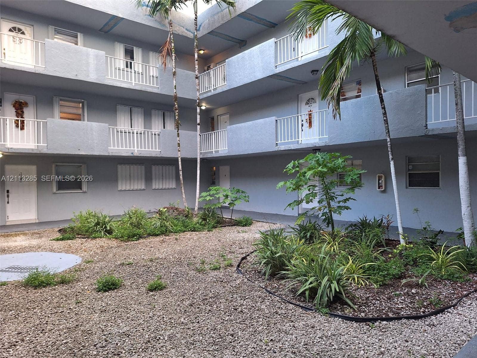 Multi-level exterior featuring white metal balcony railings, a central gravel courtyard, and mature palm trees