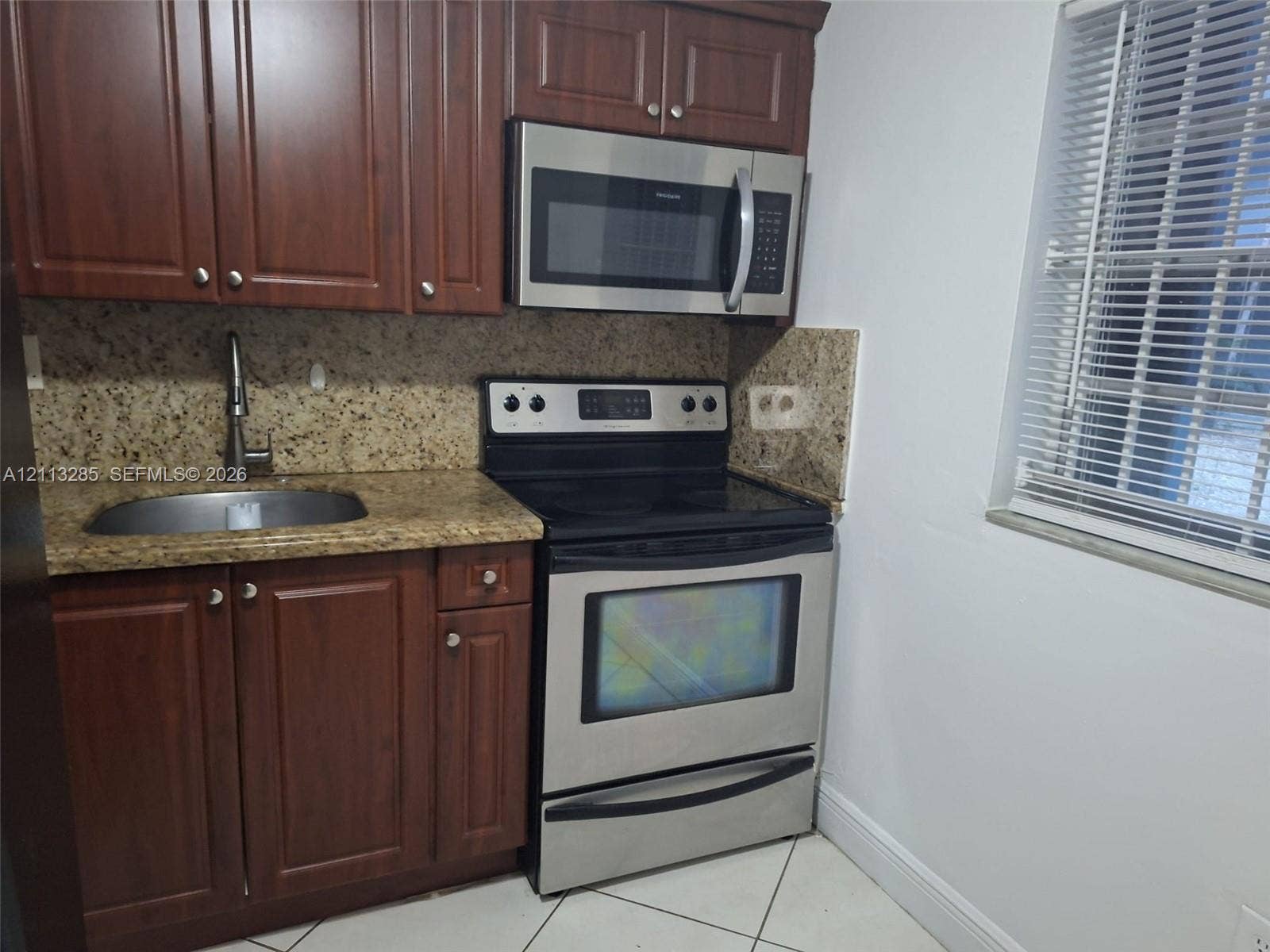 Kitchen area featuring dark-tone wood cabinetry, speckled stone countertops, stainless steel appliances, an undermount sink with a gooseneck faucet, and tile flooring
