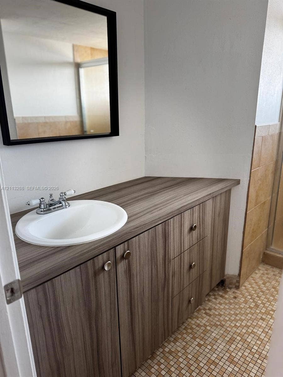Vanity featuring wood-finish cabinetry, a drop-in sink, dual-handle chrome faucet, wall-mounted mirror with a black frame, and tiled flooring