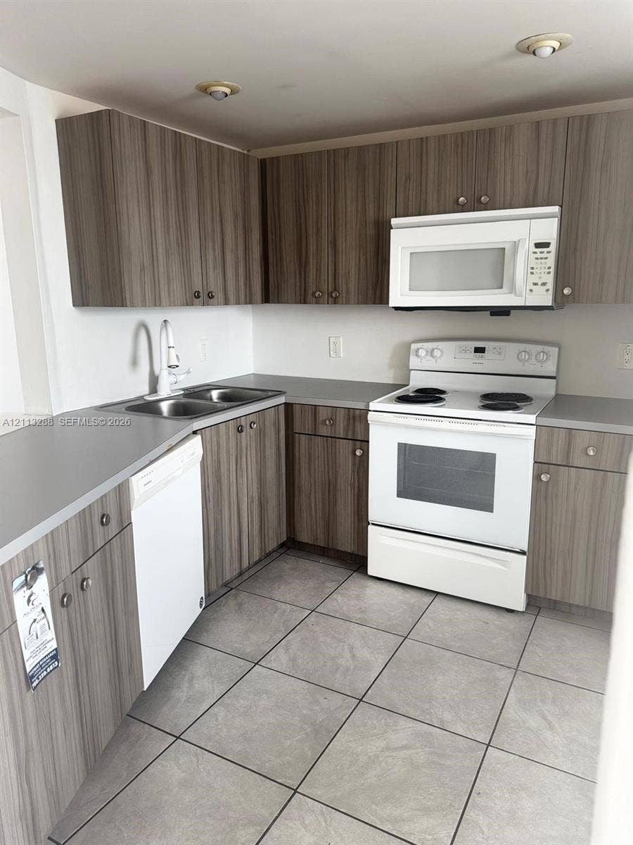 Kitchen featuring wood-finish cabinetry, neutral solid-surface countertops, large-format tile flooring, a dual-basin stainless steel sink, and white appliances