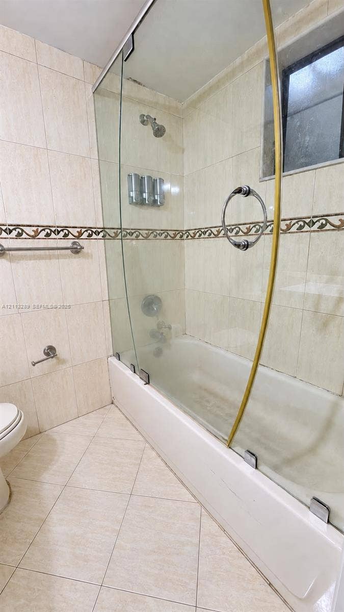 5460 W 21st Court, #208, Hialeah, FL 33016 Bathtub with a glass shower enclosure, decorative tile accent border, wall-mounted metallic fixtures, and ceramic floor tiling