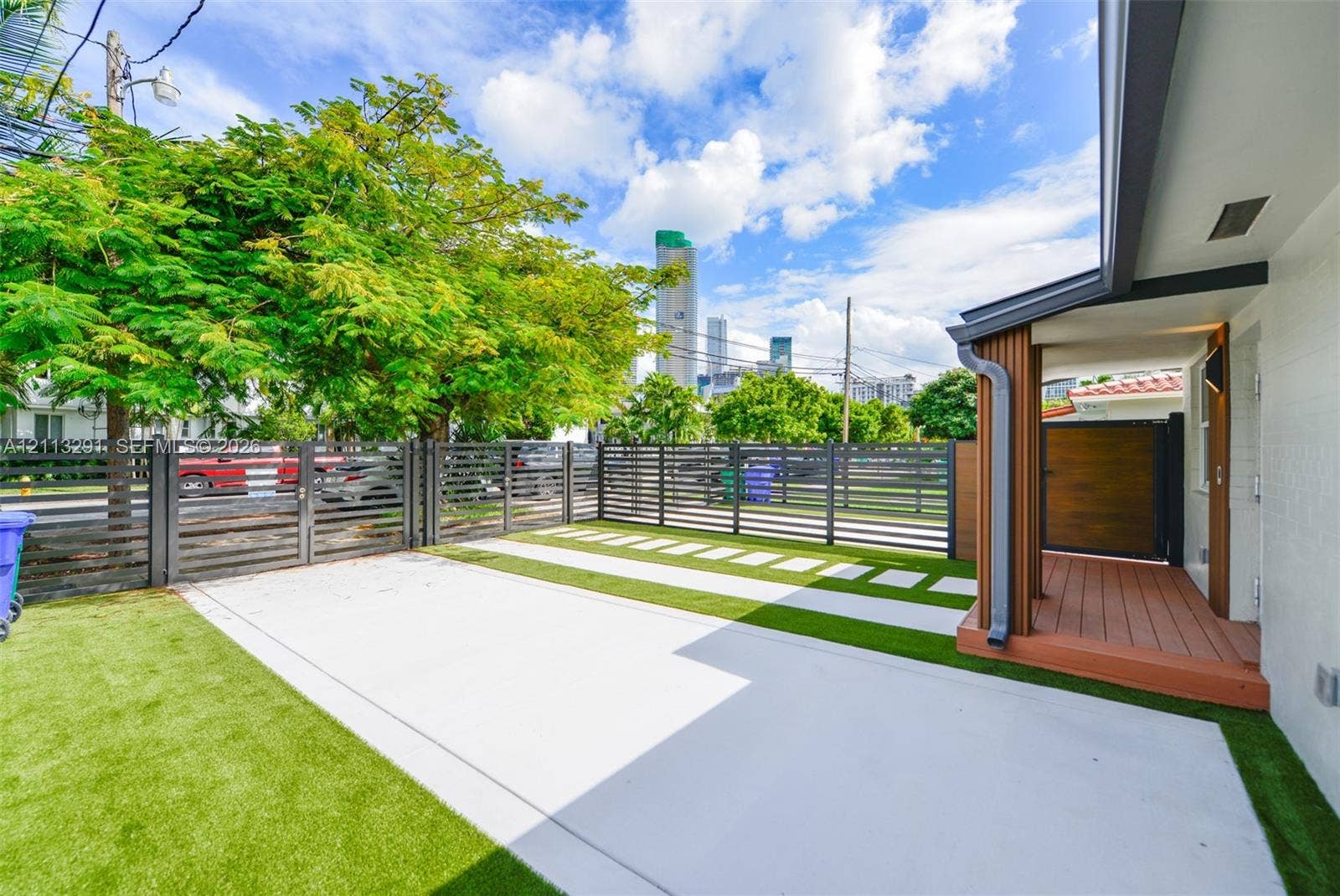 Fenced exterior space featuring concrete paving, synthetic turf, wood-finish decking, and slat-style metal fencing