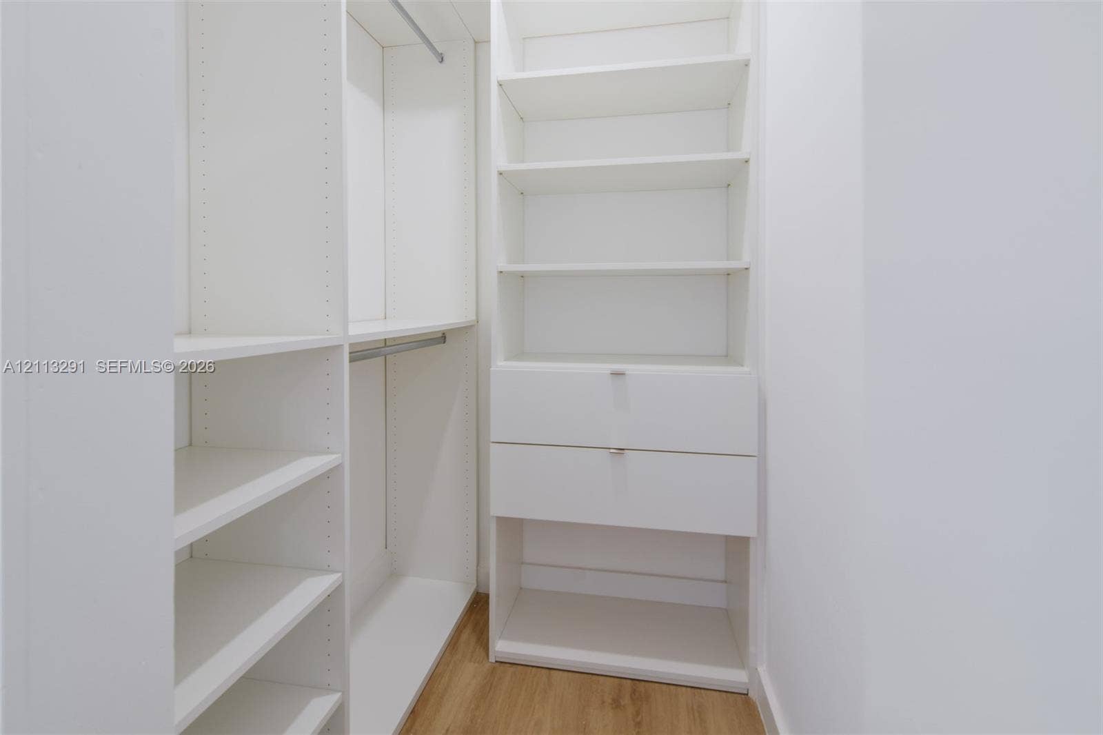 Built-in white storage system featuring adjustable shelving, integrated hanging rods, and dual drawers with silver-tone hardware over wood-finish flooring