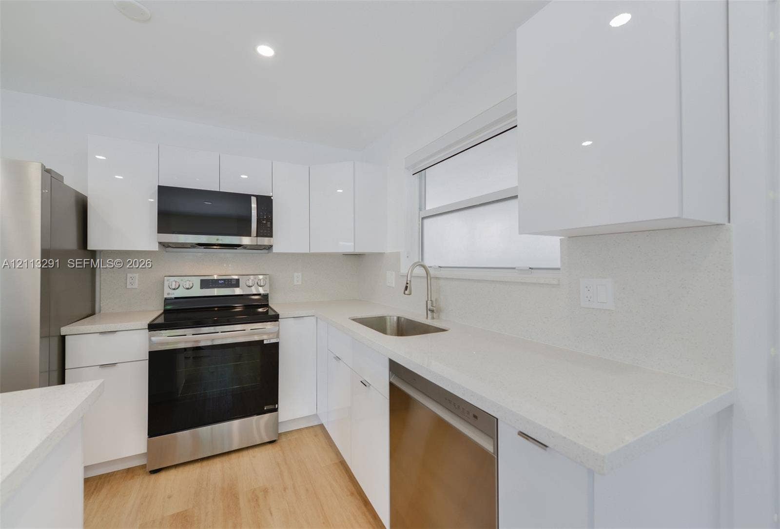 Modern kitchen featuring high-gloss white cabinetry, light-toned stone-finish countertops, stainless steel appliances, and wood-finish flooring
