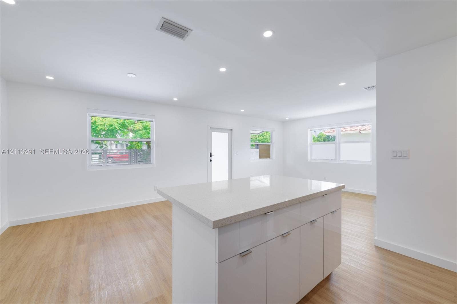 Open-concept interior featuring wood-finish flooring, recessed lighting, and a central island with a stone-composite countertop and high-gloss cabinetry