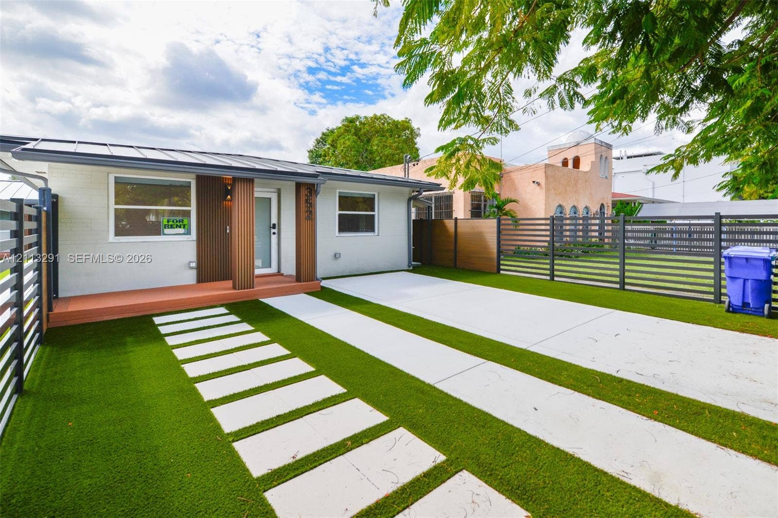 Exterior facade featuring a metal roof, synthetic turf lawn, concrete paver pathway, and horizontal privacy fencing