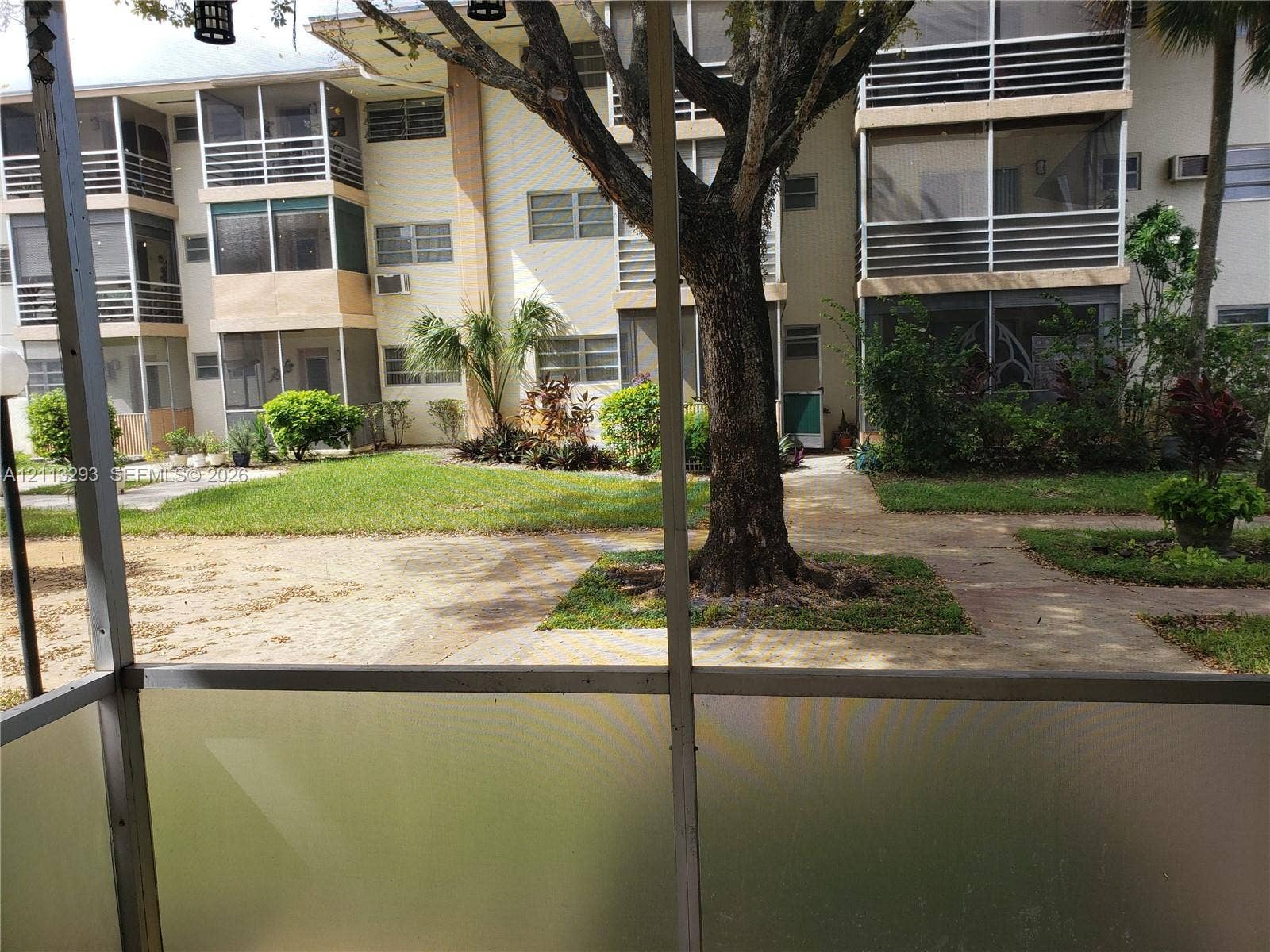Screened-in balcony featuring metal framing, overlooking a multi-story exterior with louvered windows and landscaped grounds