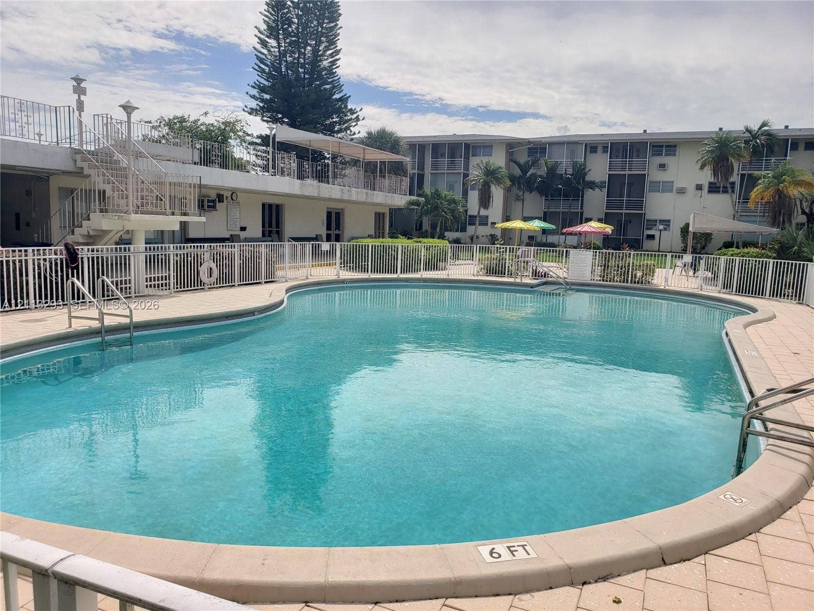 In-ground swimming pool featuring a tiled perimeter and metal ladder handrails, surrounded by a paved patio deck and enclosed by a white security fence
