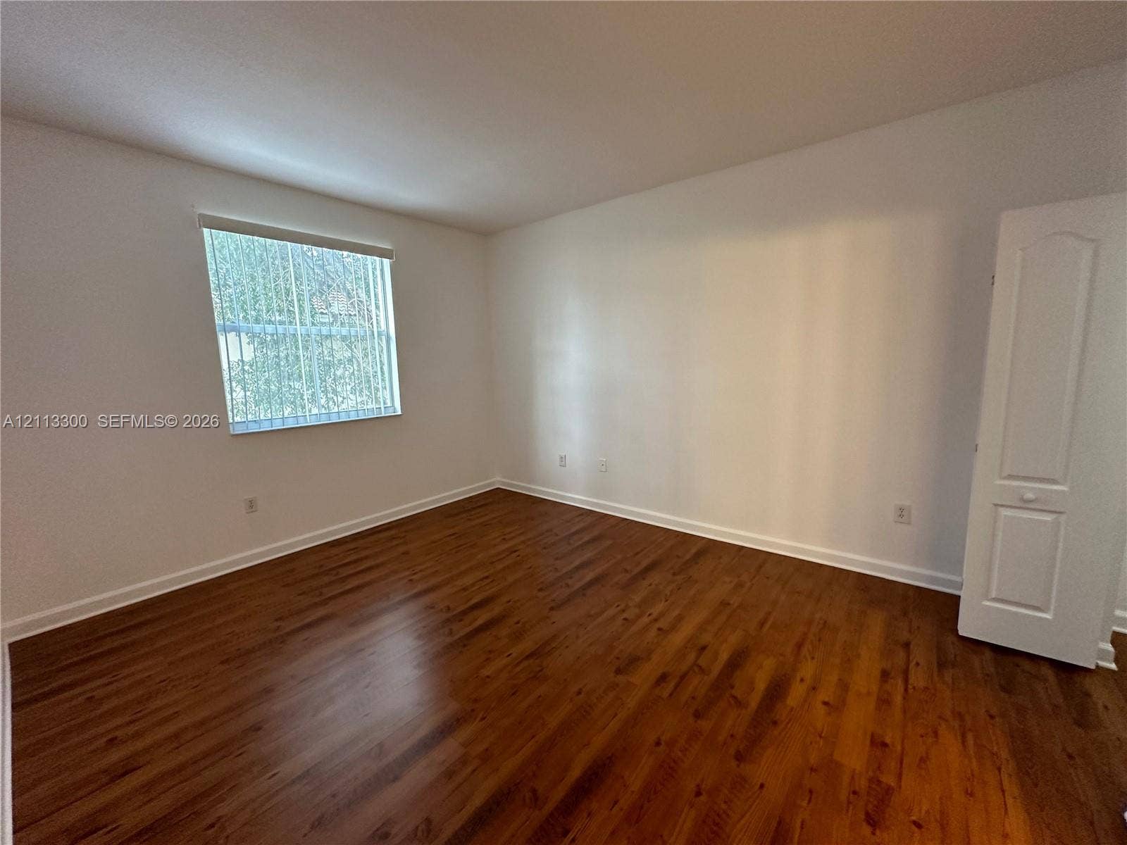 Room featuring wood-finish flooring, neutral walls, a single-panel interior door, and a window with vertical blinds