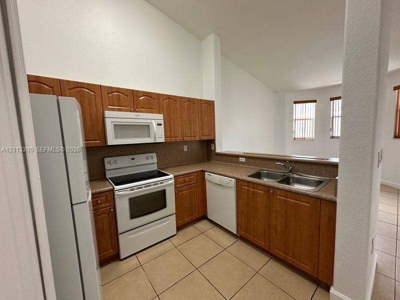 Kitchen featuring wood-finish cabinetry, neutral-toned countertops, double-basin stainless steel sink, ceramic tile flooring, and vaulted ceilings