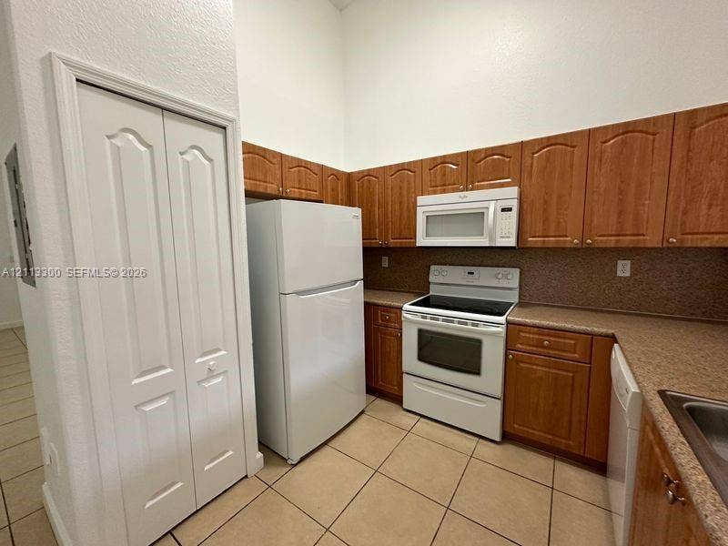 Kitchen featuring wood-finish cabinetry, white appliances, solid-surface countertops, tile flooring, and a bi-fold pantry door