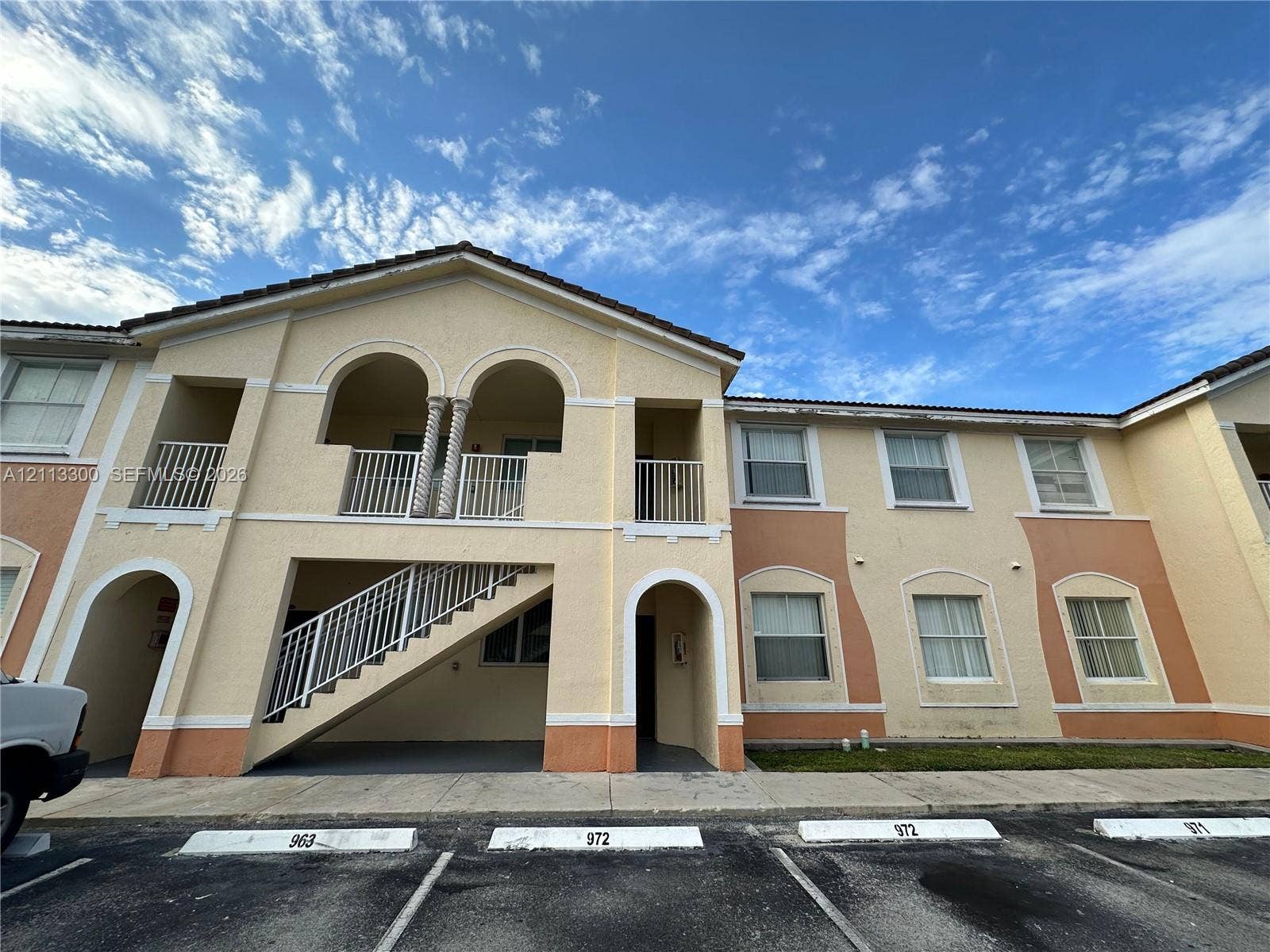 Two-story exterior featuring stucco siding, arched entryways, second-story balcony with columns, and a concrete staircase with metal railing
