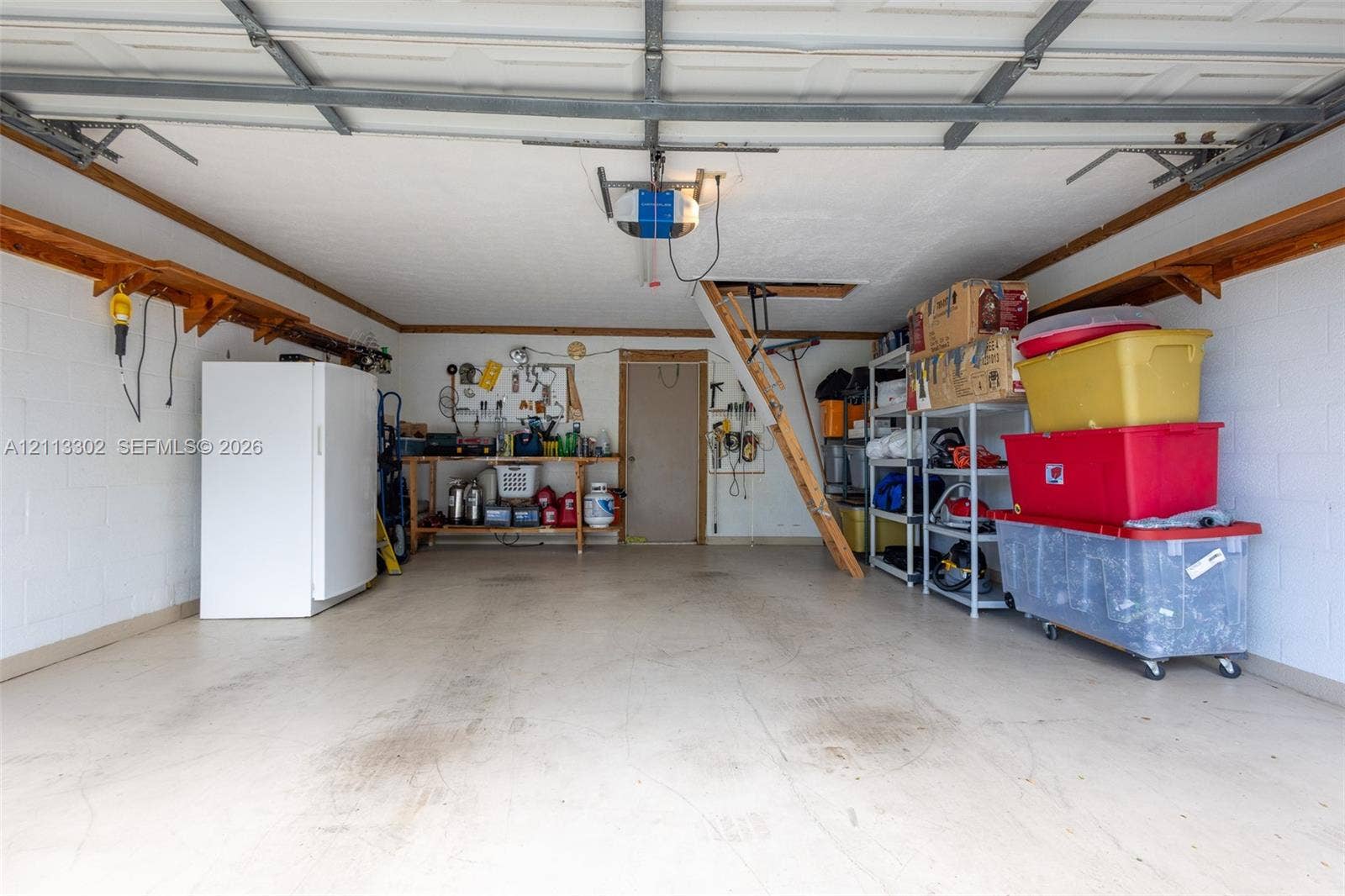 9450 Meadowood Drive, #202, Fort Pierce, FL 34951 Attached garage featuring concrete block walls, overhead door opener system, built-in wooden perimeter shelving, and pull-down attic access ladder