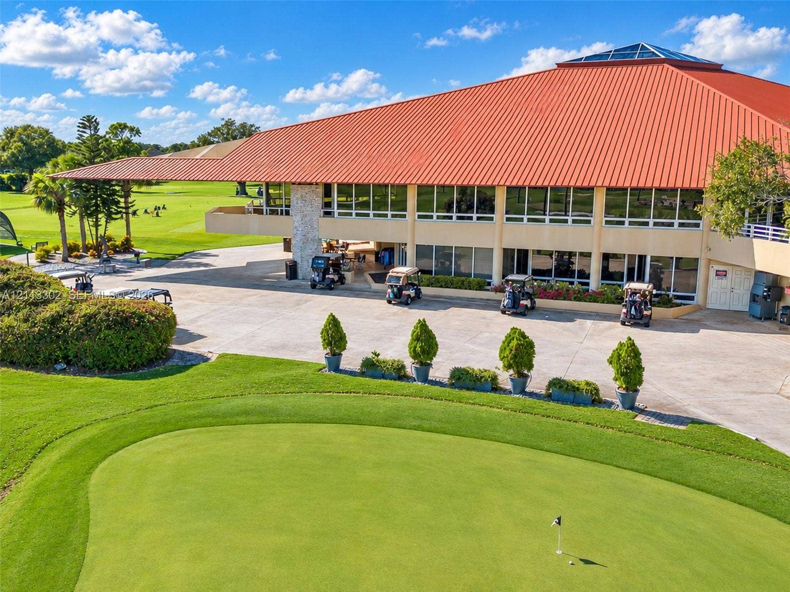 9450 Meadowood Drive, #202, Fort Pierce, FL 34951 Multi-level building featuring a prominent orange metal roof, floor-to-ceiling windows, stone pillar accents, and a concrete cart path, situated on a golf course