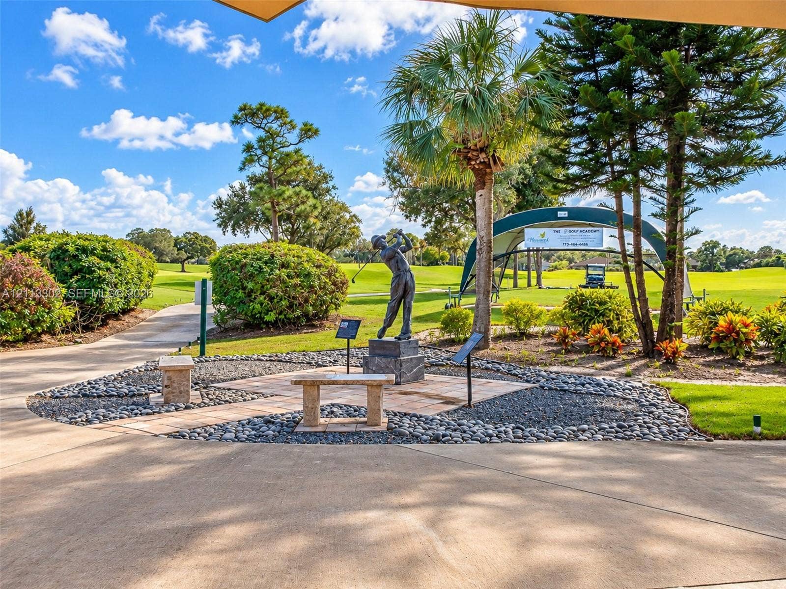 9450 Meadowood Drive, #202, Fort Pierce, FL 34951 Landscaped grounds featuring stone benches, a central bronze statue, paved pathways, mature pine and palm trees, and golf course frontage