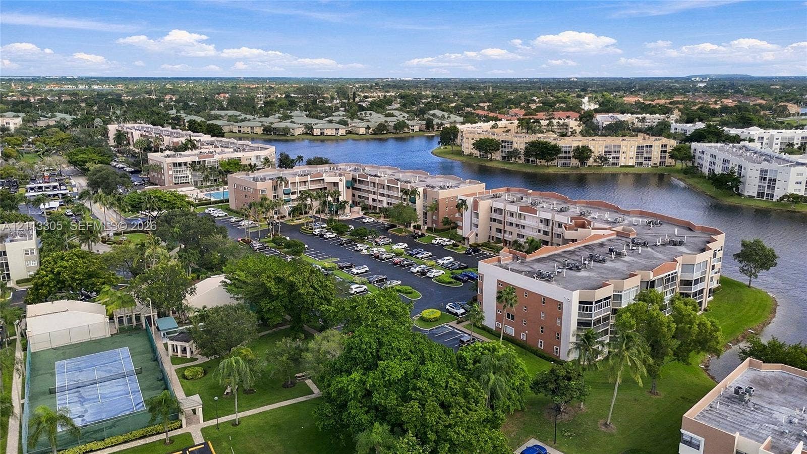 Aerial perspective of a multi-story residential complex featuring waterfront positioning, an enclosed tennis court, surface parking, and manicured landscaping