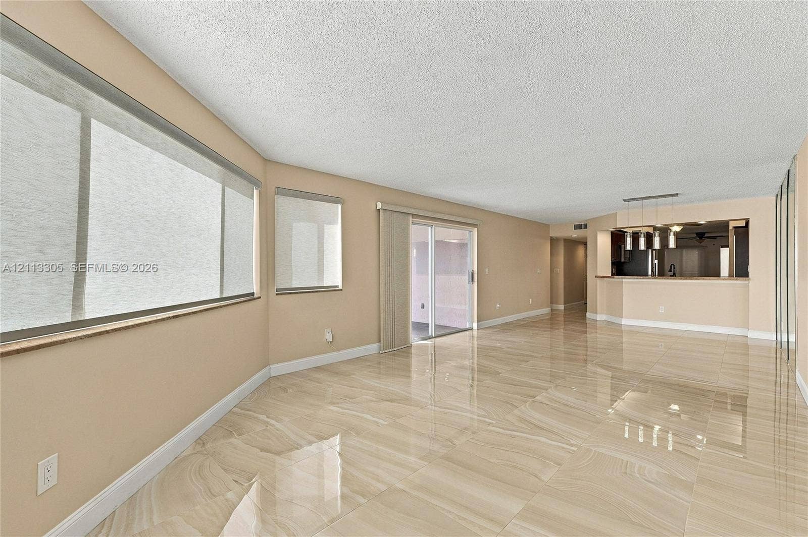 Large-format polished tile flooring, sliding glass door with vertical blinds, expansive picture window, and pass-through kitchen counter with multi-pendant lighting