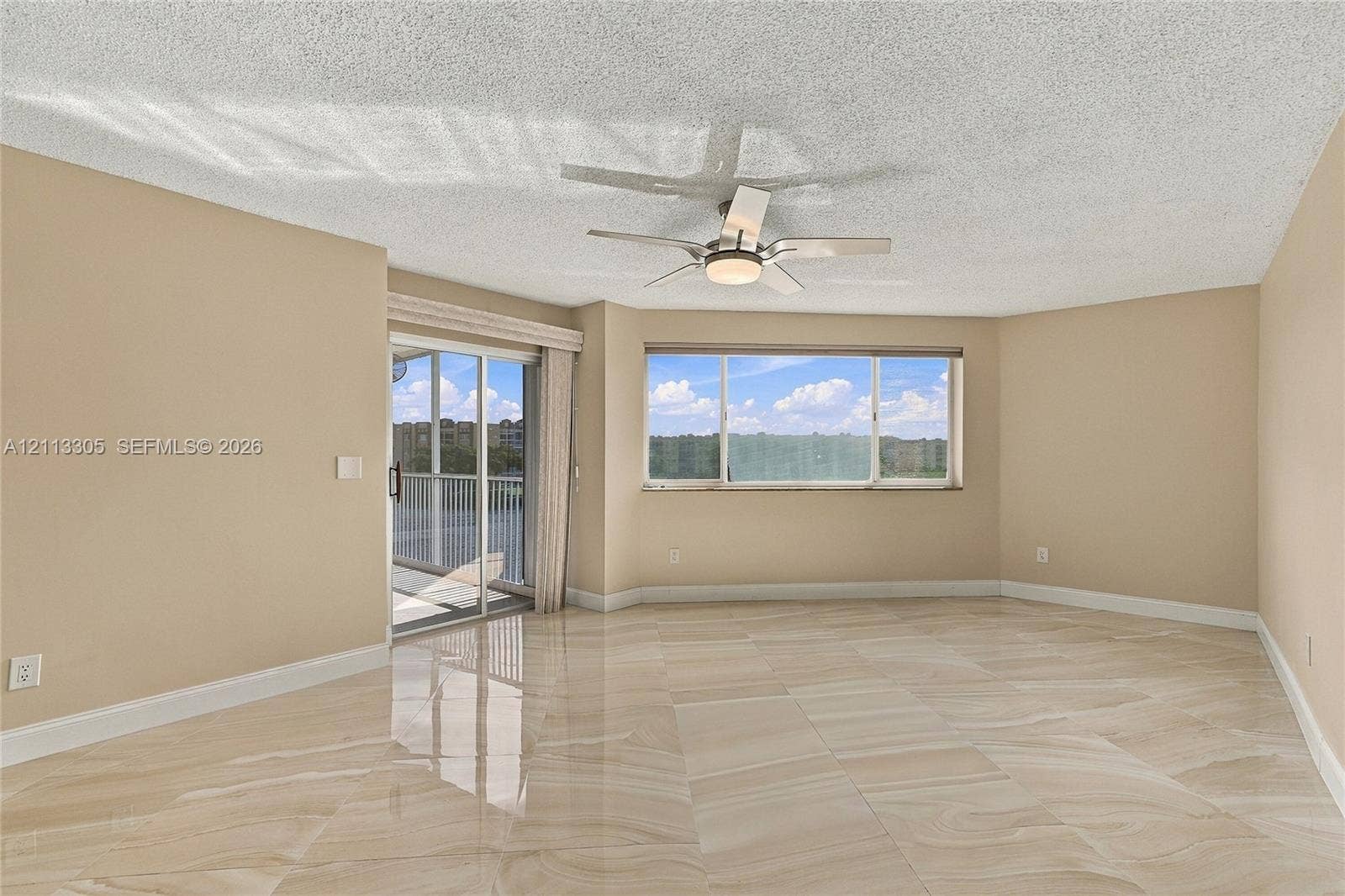 Neutral-toned interior featuring large-format polished tile flooring, ceiling fan with integrated lighting, horizontal sliding window, and sliding glass door with external balcony access