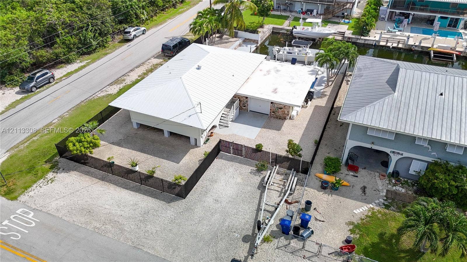 Elevated structure featuring a white metal roof, stone-veneer exterior accents, a gated gravel perimeter, and private canal bulkhead