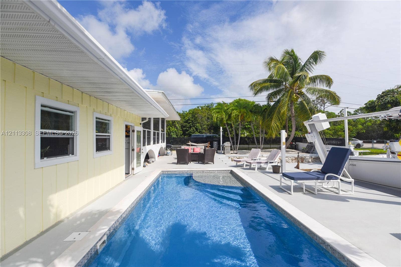 Rectangular in-ground swimming pool with light-toned paved decking, light yellow vertical-siding exterior, and integrated boat lift