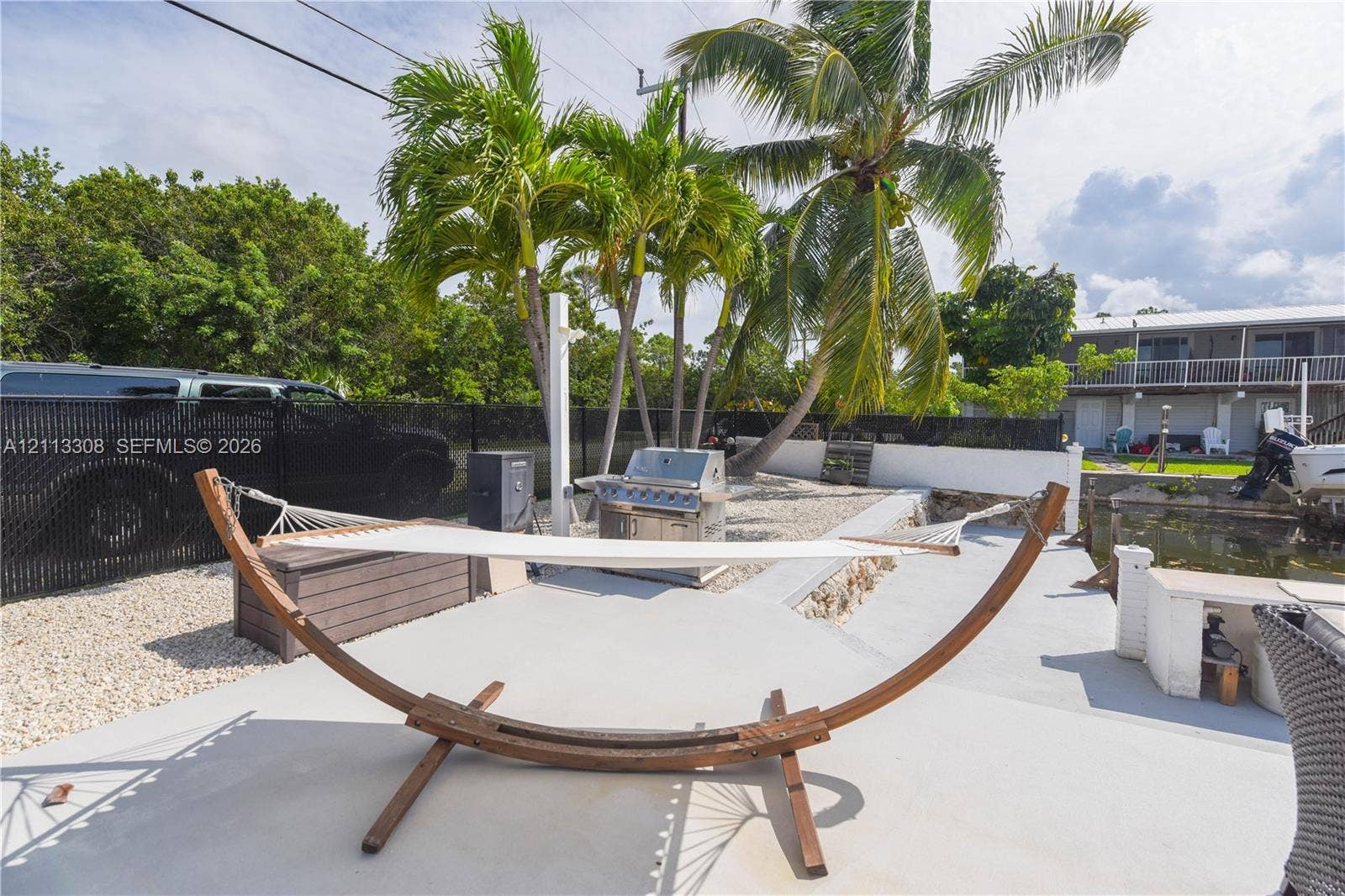 Outdoor area featuring a concrete patio, built-in stainless steel grill, stone-filled ground cover, perimeter black metal fencing, and palm trees