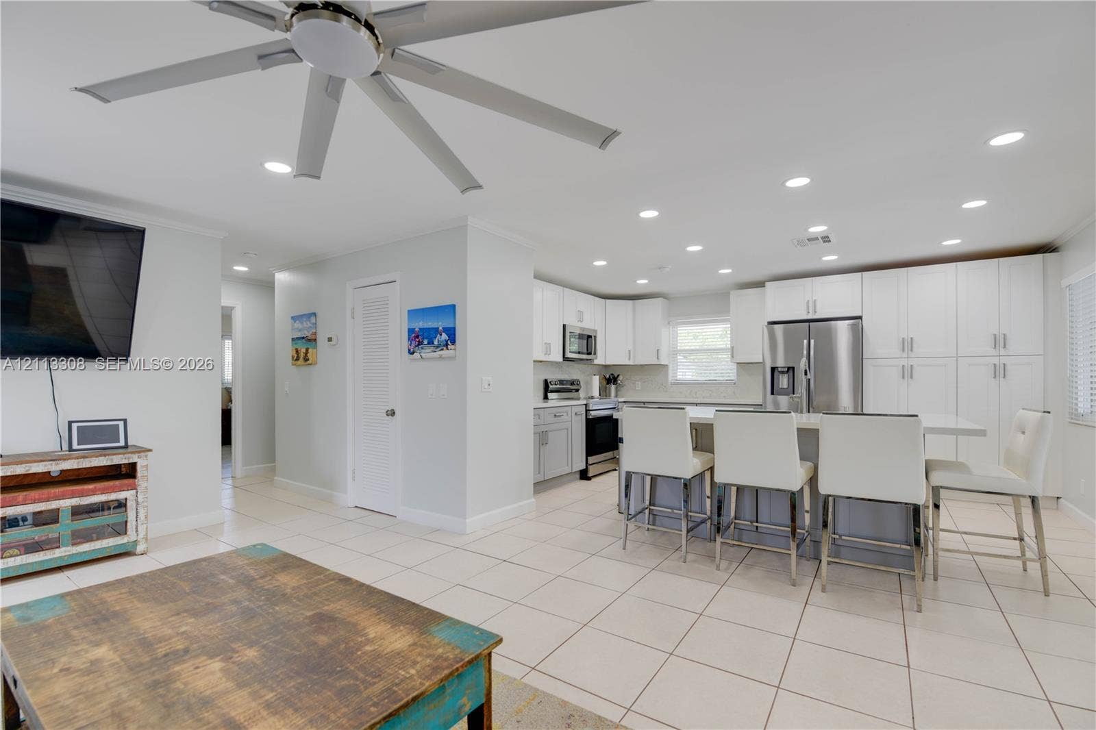 Open-concept kitchen featuring white shaker-style cabinetry, stainless steel appliances, quartz-surface breakfast bar, recessed lighting, and large-format tile flooring