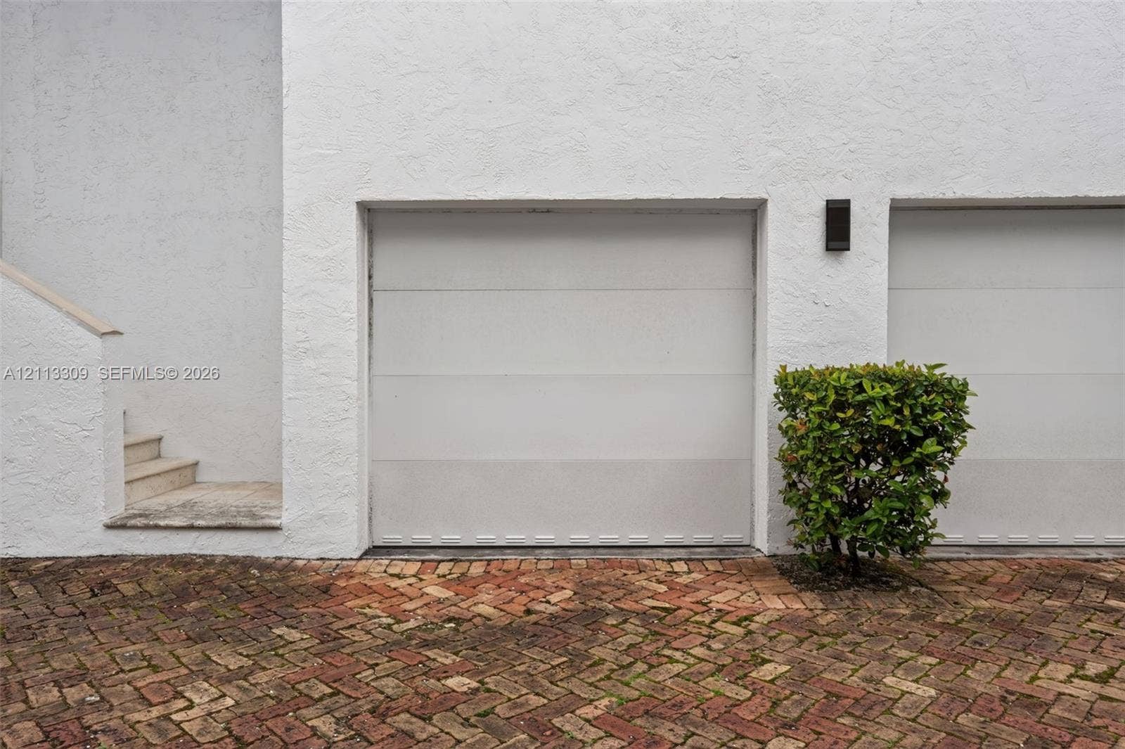 3082 Bird Avenue, #6, Miami, FL 33133 Multi-car garage featuring white paneled doors, textured stucco exterior, exterior wall-mounted lighting, and a herringbone-patterned brick paver driveway