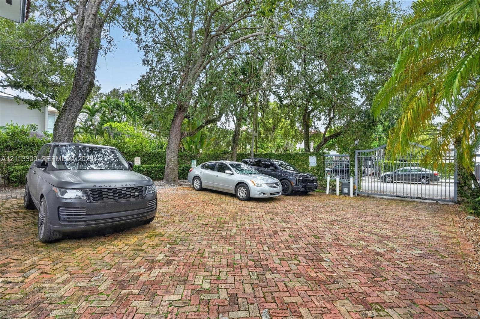 3082 Bird Avenue, #6, Miami, FL 33133 Brick-paved parking area featuring mature trees, perimeter hedging, and a metal security gate