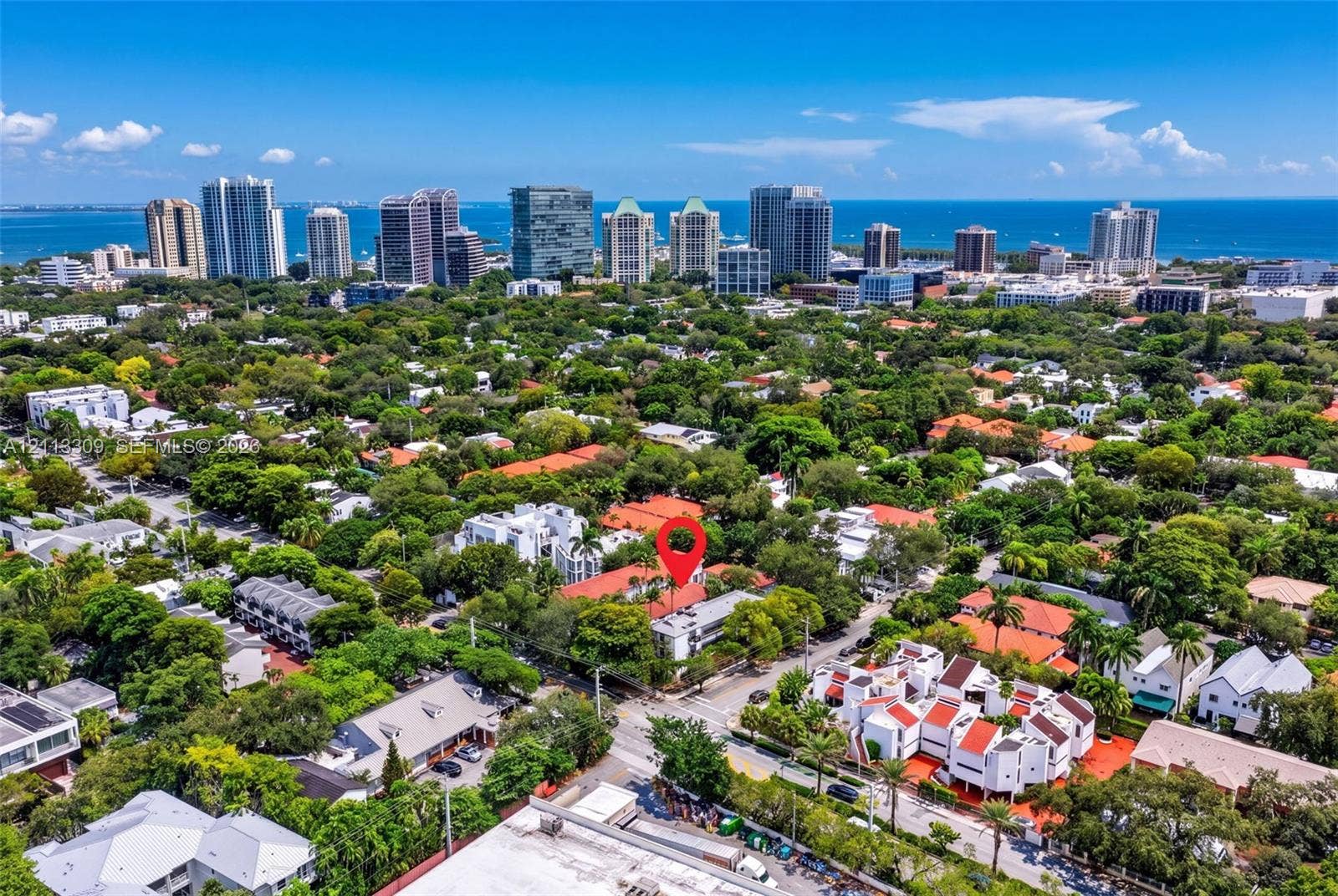 3082 Bird Avenue, #6, Miami, FL 33133 Aerial perspective highlighting a multi-unit residential structure featuring white exterior masonry and terracotta-style roof tiles, situated within a dense canopy of mature trees with proximity to a coastal skyline