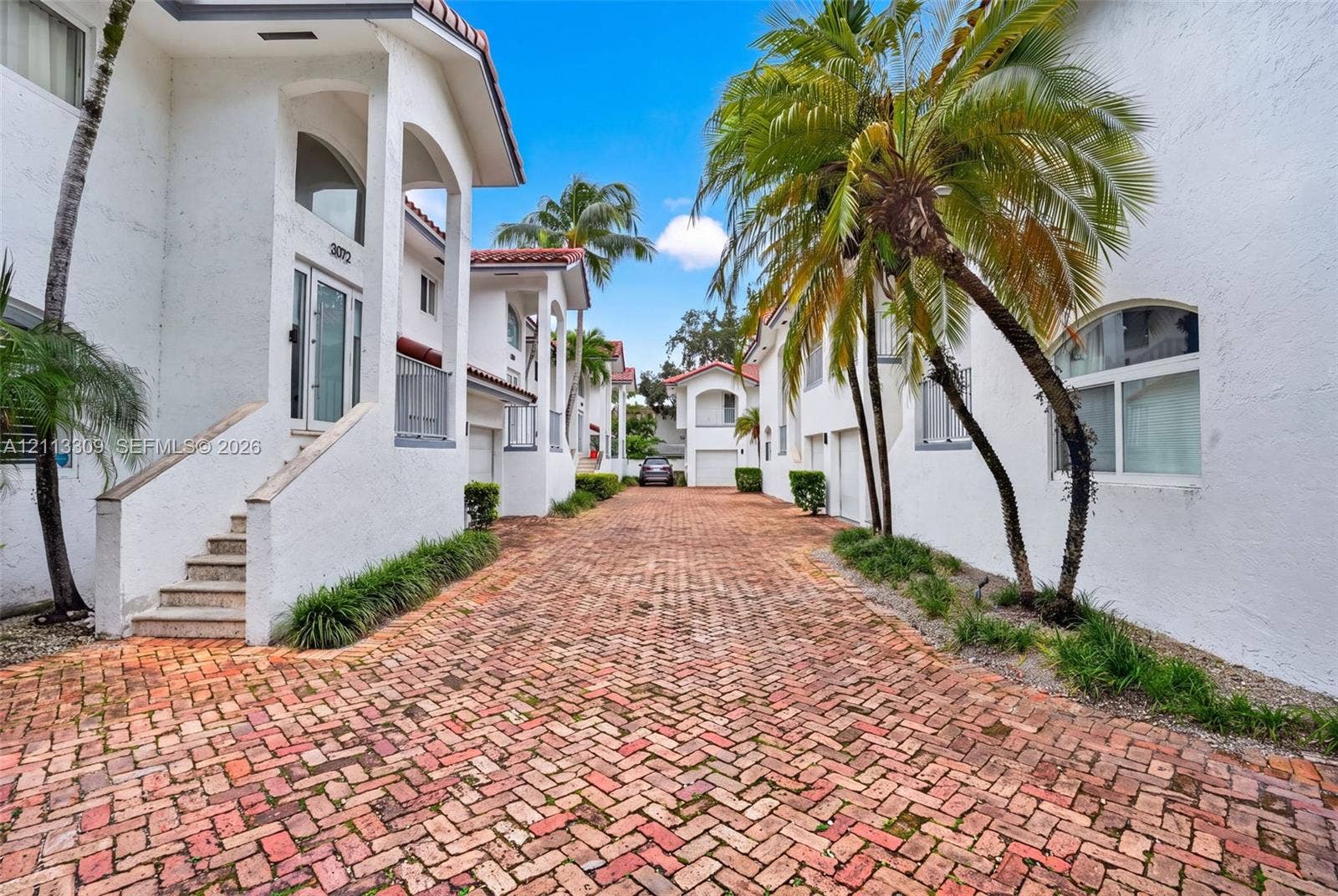 3082 Bird Avenue, #6, Miami, FL 33133 Stucco exterior featuring arched architectural detailing, a herringbone-patterned brick paver motor court, and mature palm landscaping