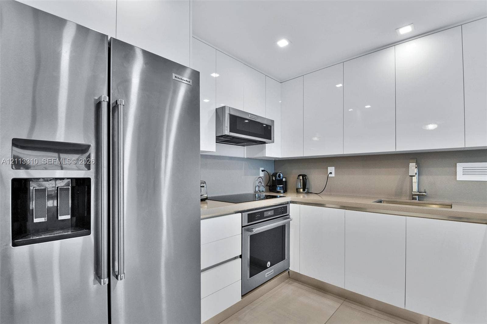 Kitchen featuring high-gloss white cabinetry, solid surface countertops, recessed lighting, and stainless steel appliances including a built-in oven, over-the-range microwave, and side-by-side refrigerator
