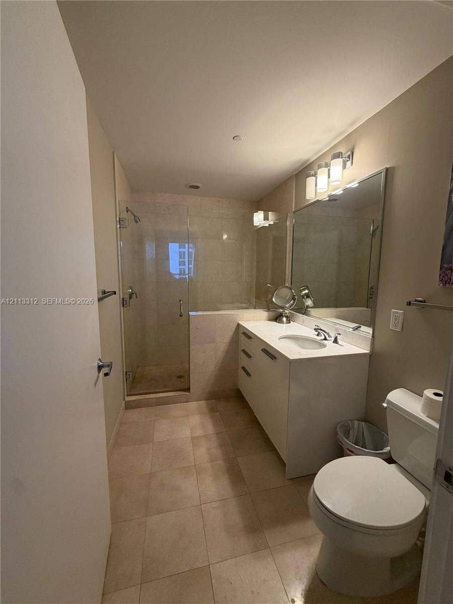 Bathroom featuring a white vanity with an integrated basin, a large wall-mounted mirror, neutral tile flooring, and a glass-enclosed walk-in shower with tile surround