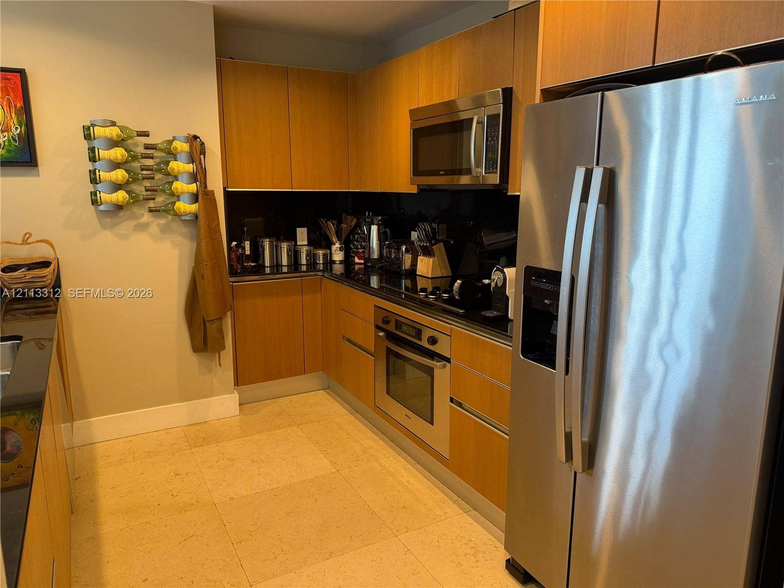 Kitchen featuring wood-veneer flat-panel cabinetry, black countertops, stainless steel wall-mounted oven and microwave, and light-toned stone-finish tile flooring