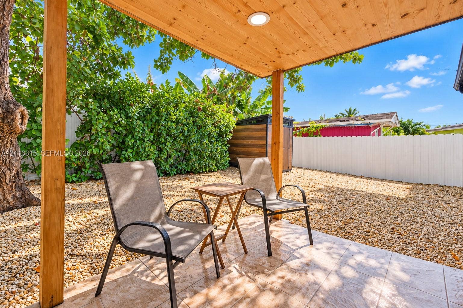 Covered patio featuring a light wood-toned tongue-and-groove ceiling with recessed lighting, tiled flooring, and a gravel-surfaced yard enclosed by a white vinyl privacy fence