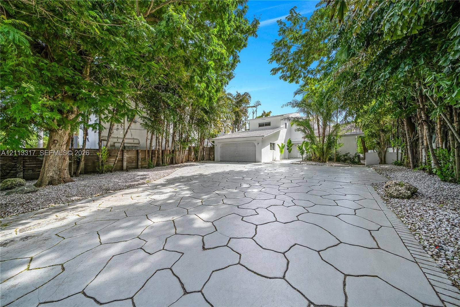 Extensive paver driveway featuring a decorative flagstone pattern, mature perimeter trees, stone-accented landscaping, and an attached garage