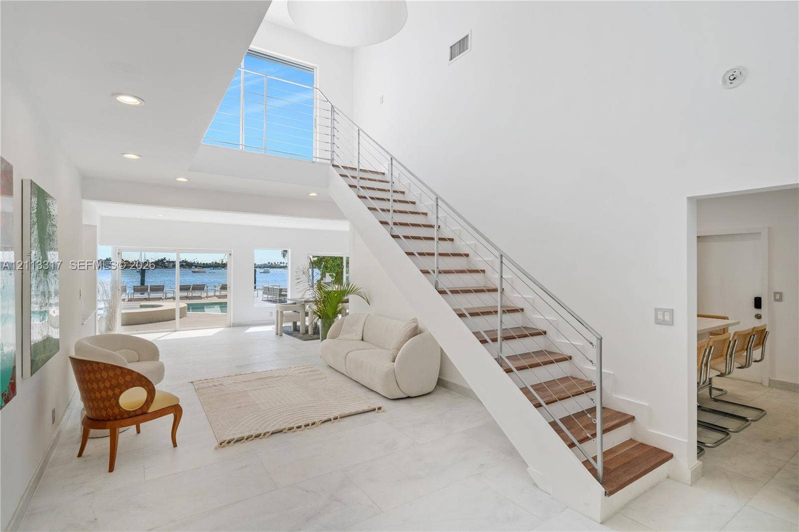 Open-concept interior featuring high ceilings, open-riser staircase with stainless steel cable railing and wood-finish treads, large-format tile flooring, and floor-to-ceiling windows