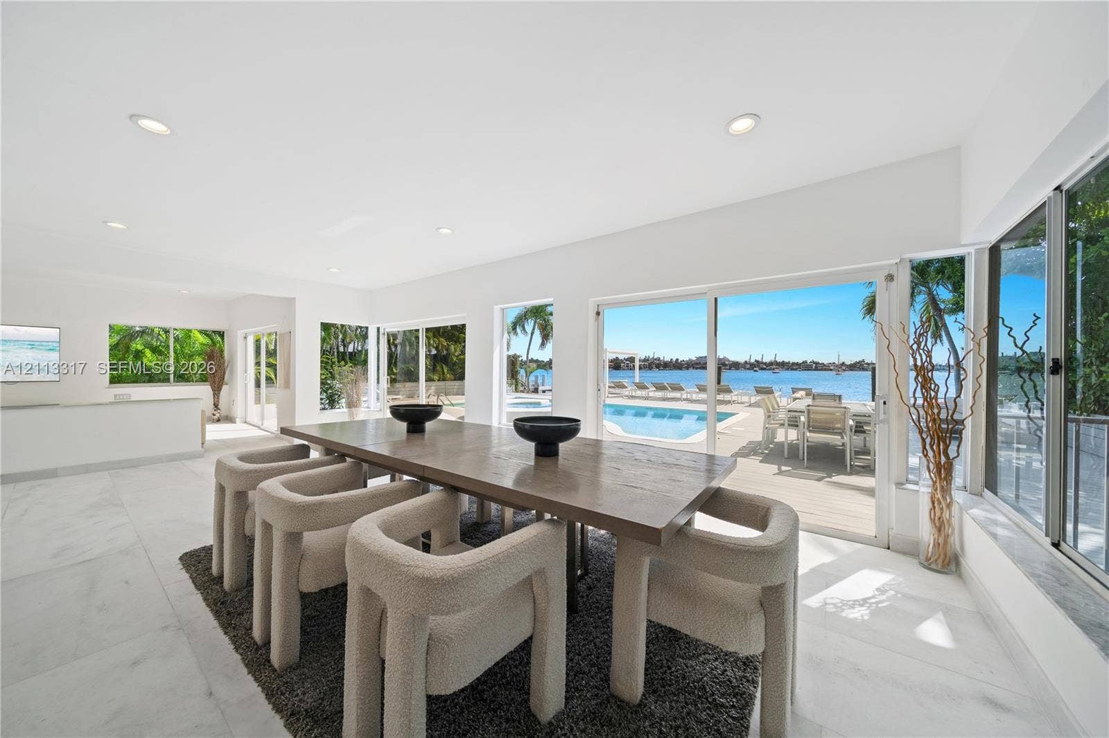 Open-concept interior featuring large-format tile flooring, recessed lighting, and expansive floor-to-ceiling glass sliding doors opening to an outdoor pool and waterfront