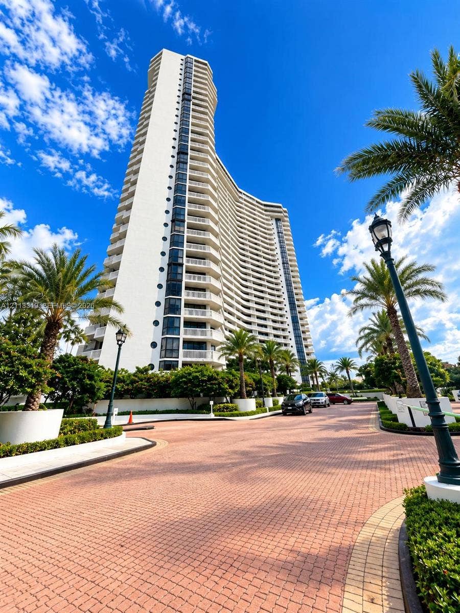 High-rise building featuring a white facade, curvilinear architectural design, stacked private balconies, and a paved brick entrance drive lined with mature palm trees and decorative lighting fixtures