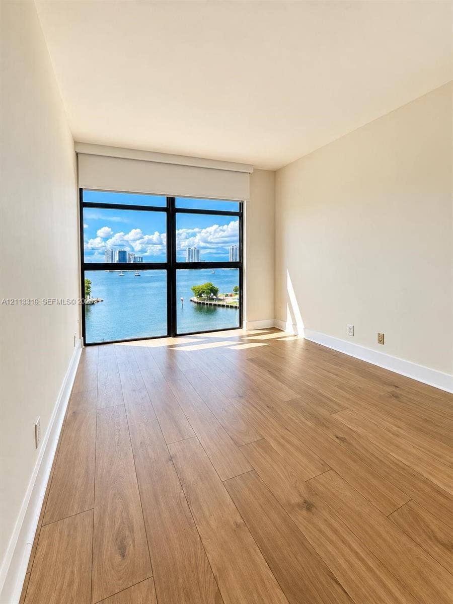 Floor-to-ceiling windows featuring water and skyline scenery, wood-finish flooring, and a window shade